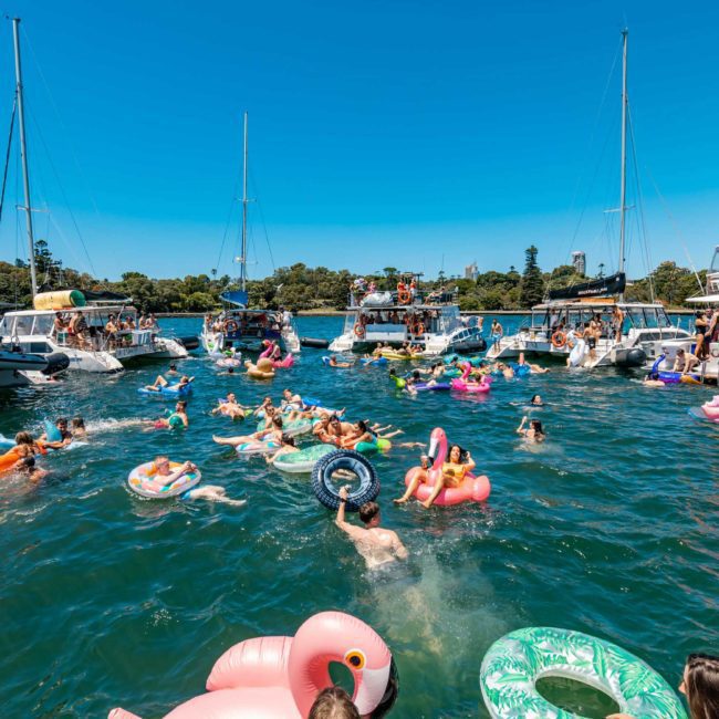 Dozens of people enjoy a sunny day in the water, surrounded by boats and colorful inflatable pool floats, with trees visible in the background—a perfect setting for a Sydney boat party hire.
