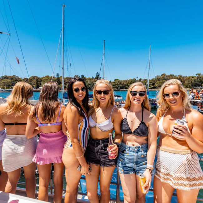 A group of women in swimsuits and summer outfits stand on a boat deck, holding drinks, with additional people and other boats visible in the background, enjoying a vibrant Catamaran party Sydney.