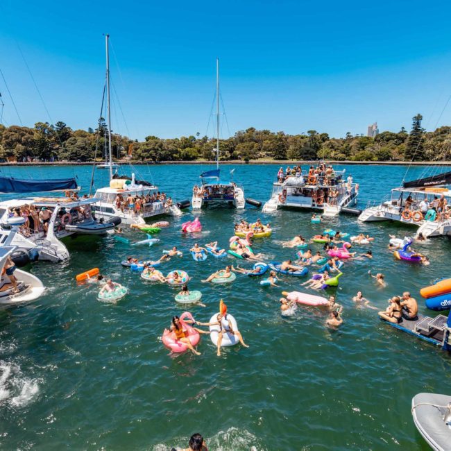 A group of people are enjoying a sunny day on a lake, floating on inflatable rafts and swimming near several anchored boats. Trees and buildings are visible in the background, hinting at the perfect setting for Corporate boat events Sydney or even DJ boat hire Sydney.