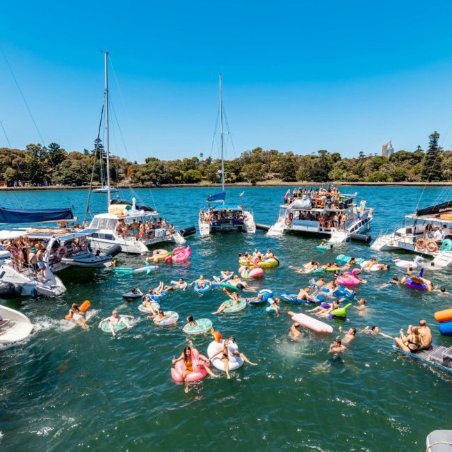 People enjoying a sunny day on a lake with boats and inflatable floats, surrounded by trees in the background, reminiscent of a catamaran party on Sydney Harbour.