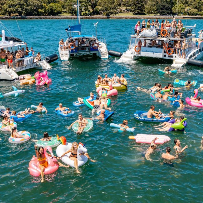 A crowded lake scene with people on inflatable floats and boats, enjoying a sunny day. The water is filled with various brightly colored inflatables, creating a lively atmosphere. It's reminiscent of a Sydney boat party hire event, with the serene backdrop of trees adding to the festive mood.