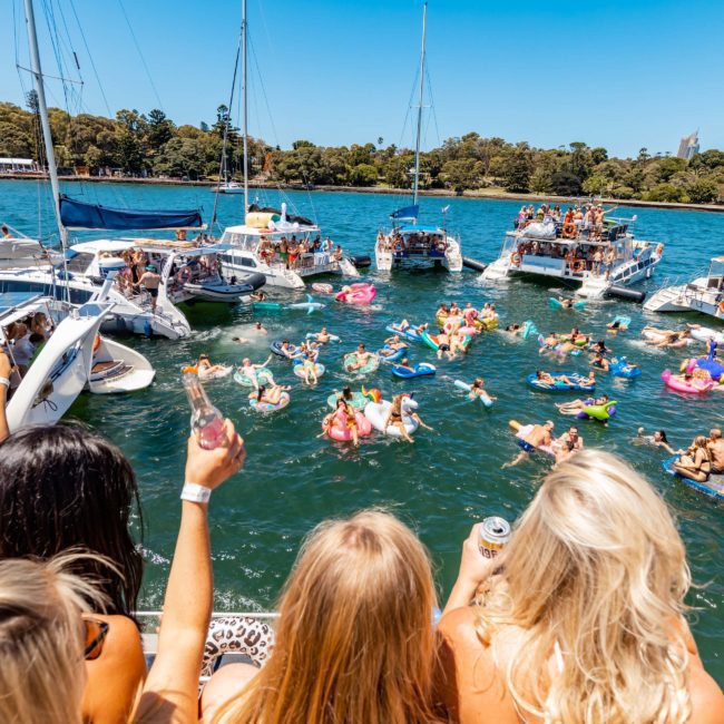 People are on boats and inflatables on the water with several onlookers in the foreground enjoying a sunny day, perfect for a Sydney boat party hire or a private yacht charter Sydney Harbour adventure.