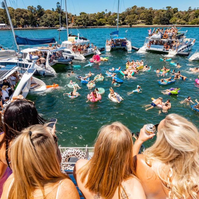 People are enjoying a sunny day on a group of boats and inflatables in a lively gathering on Sydney Harbour. The scene is festive with numerous attendees and various types of boats, including private yacht charters and catamaran parties, creating an unforgettable experience on the water.
