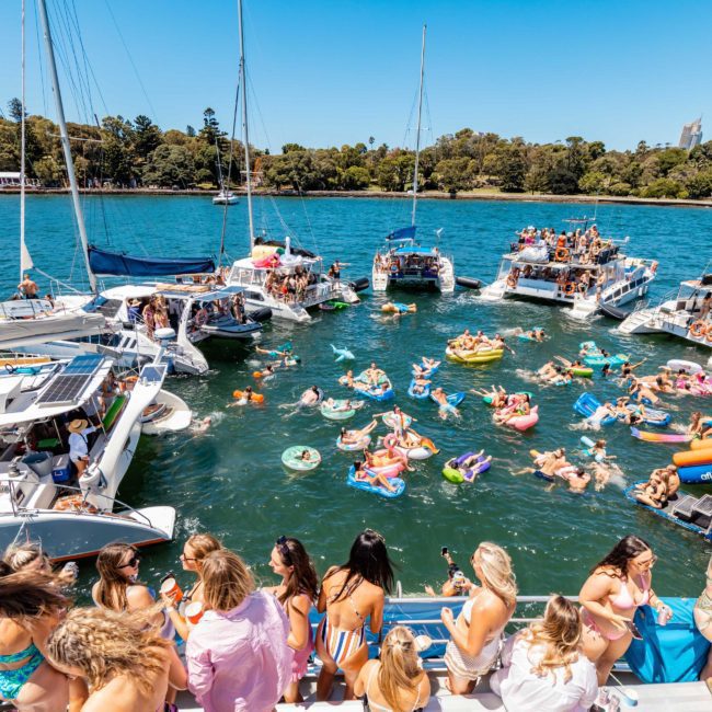 A large group of people enjoy a sunny day on the water, swimming and lounging in inflatable floats, while several boats are anchored nearby. The shoreline with trees is visible in the background, setting a perfect scene for a Sydney boat party hire or a DJ boat hire Sydney event.