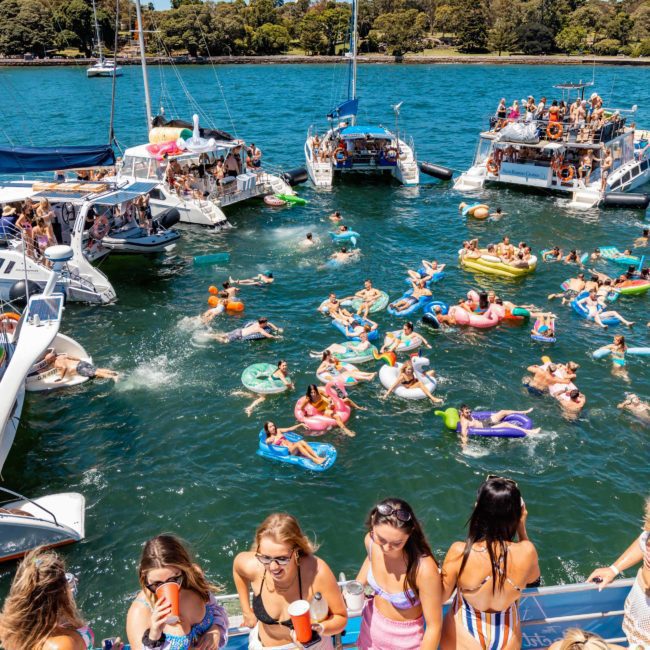 A group of people enjoy a summer boat party on a luxury yacht hire in Sydney Harbour, with many floating in the water on inflatable rafts and others socializing on nearby boats under a clear sky.