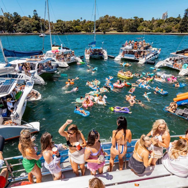 People partying on yachts and floating in the water on a sunny day, surrounded by other boats and trees in the background. It's the perfect scene for a catamaran party Sydney, where everyone is enjoying their time under the sun.