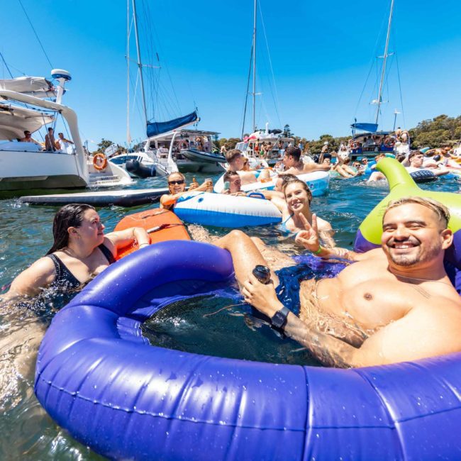 A group of people enjoying a sunny day in a crowded marina, floating on colorful inflatables and surrounded by boats, highlights the vibrant atmosphere you get with DJ boat hire Sydney.