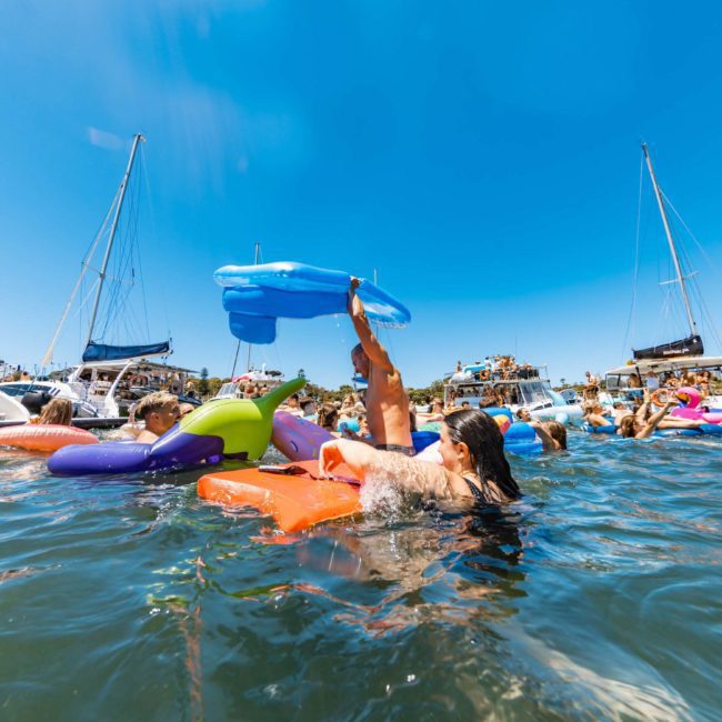 People enjoying a sunny day in the water, floating on various inflatable devices near anchored boats and yachts, creating an atmosphere reminiscent of a Sydney boat party hire.