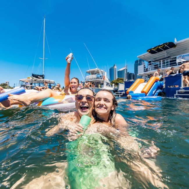 A group of people enjoy a sunny day swimming and playing on inflatables in a body of water, with boats moored in the background. They relish the excitement of a private yacht charter on Sydney Harbour, making the most of their aquatic adventure.
