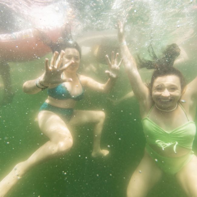 Two women in swimsuits are underwater, smiling and waving toward the camera. The water is greenish, and their hair floats around them. Other swimmers are visible in the background, enjoying a sunny day reminiscent of lively Sydney boat party hire events.