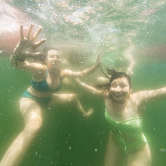 Two people in swimsuits are underwater in a green-tinged body of water, smiling and reaching towards the camera, embodying the joyful spirit often found at private yacht charters on Sydney Harbour.