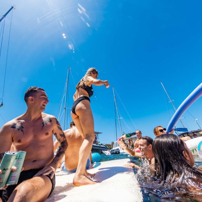 A group of people enjoying a sunny day on a boat in the water, with some individuals floating nearby on pool noodles. One person is standing on the boat, while others swim and laugh around. It looks like they opted for a Private yacht charter Sydney Harbour, making their day even more special.