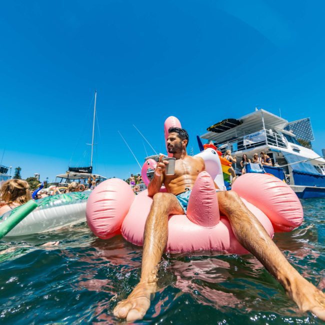 A man lounges on a pink inflatable flamingo in a body of water, surrounded by other people on inflatables and boats. City buildings and a blue sky are visible in the background, ideal for corporate boat events Sydney or DJ boat hire Sydney.
