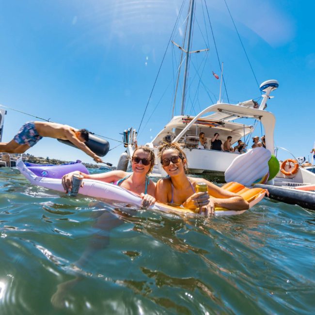 Two people in swimsuits float on inflatable loungers in clear water, smiling on a sunny day. Behind them, others enjoy a lively Sydney boat party hire on luxurious yachts and boats.
