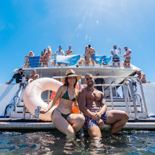 People on a boat enjoy a sunny day; a man and woman in swimwear pose near the water holding a life ring, making the most of their DJ boat hire Sydney experience.