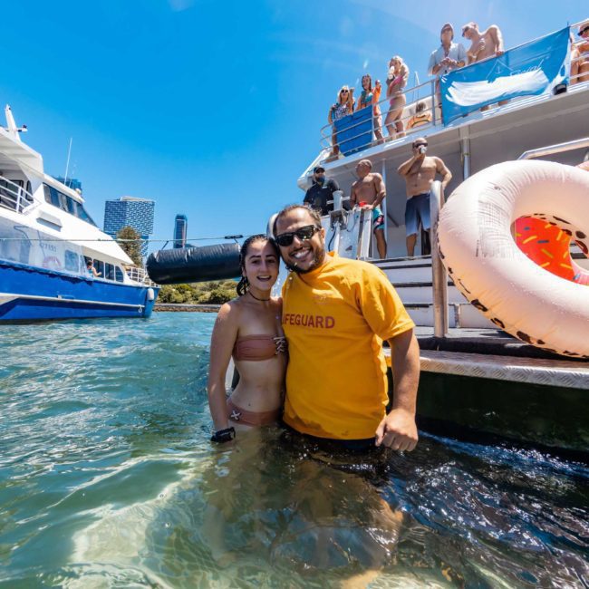 A lifeguard and a woman stand in the water by a boat on a sunny day. People are on the boat, and various inflatable items are visible. It looks like the perfect setting for a private yacht charter Sydney Harbour experience or even a catamaran party Sydney event.