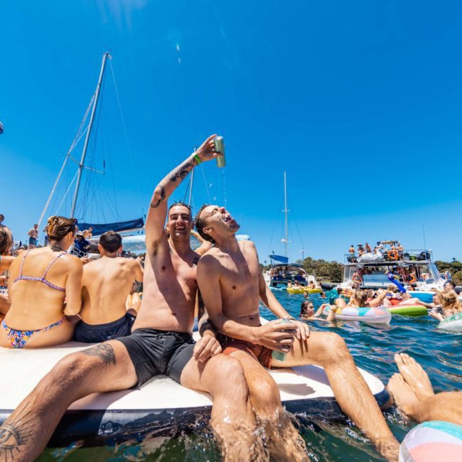 People in swimsuits on a dock and boats, enjoying a sunny day at the beach. Two men in the center are pouring a drink, with others swimming and relaxing nearby during a private yacht charter Sydney Harbour.