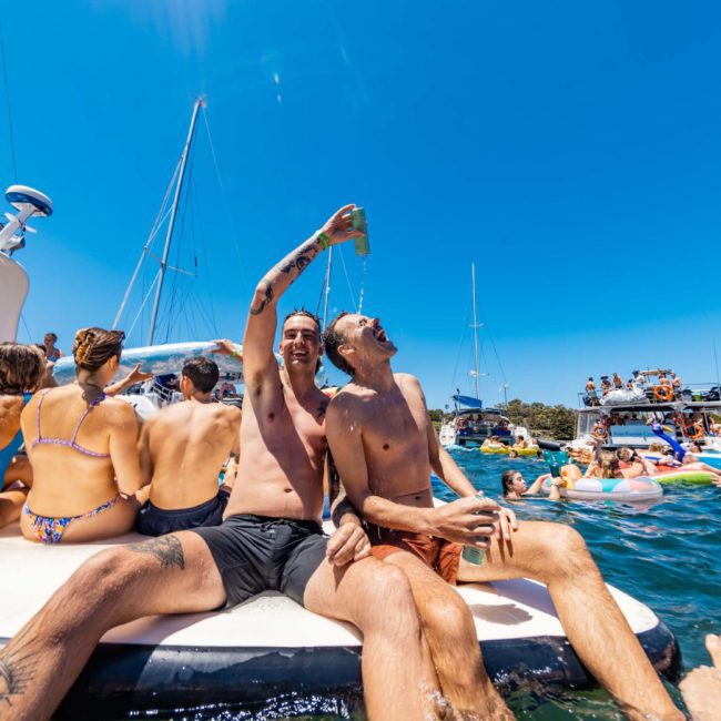 People enjoy a sunny day on boats and floaties in a body of water. Two men on a floating platform pour a drink. Other individuals relax and socialize around them under the clear blue sky. It's an idyllic scene, perfect for a luxury yacht hire in Sydney or even an exclusive catamaran party.