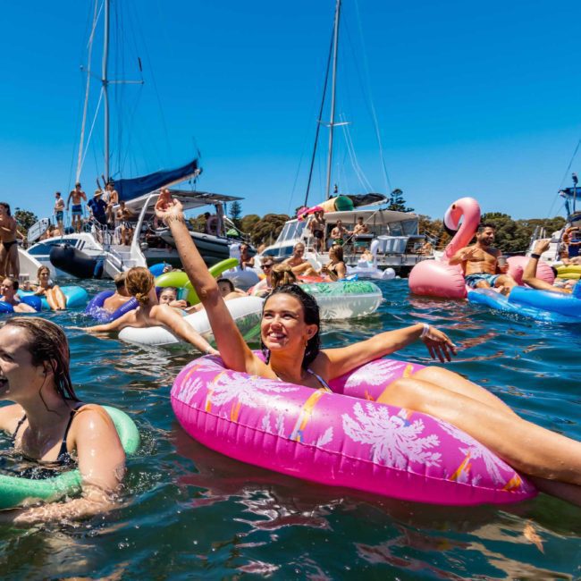 People enjoy a sunny day on the water with various inflatables and boats. A woman lounges on a pink float, and others are seen swimming and socializing around her, highlighting the lively atmosphere perfect for private yacht charter Sydney Harbour.
