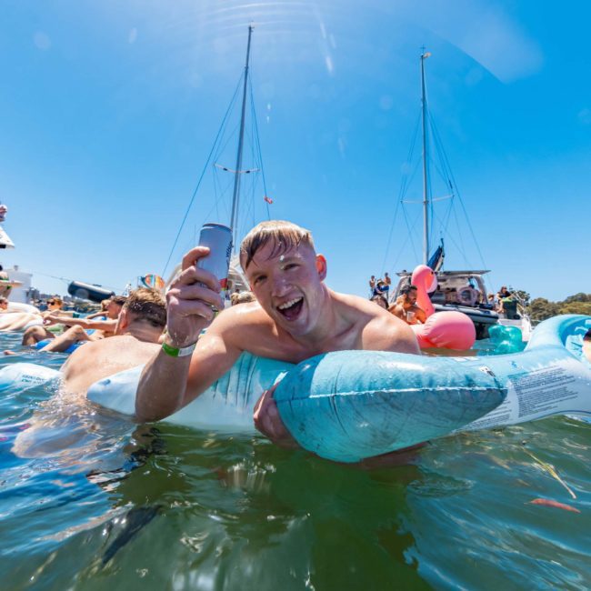 A man in a blue inflatable float is holding up a can and smiling in a body of water. There are boats and other people on floats in the background, enjoying what looks like an incredible Sydney boat party hire.