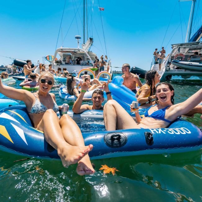 A group of people are enjoying themselves on inflatable floats in the water, with several boats in the background and a clear blue sky above. Some individuals are raising their arms and holding drinks, making it a perfect scene for a private yacht charter on Sydney Harbour.