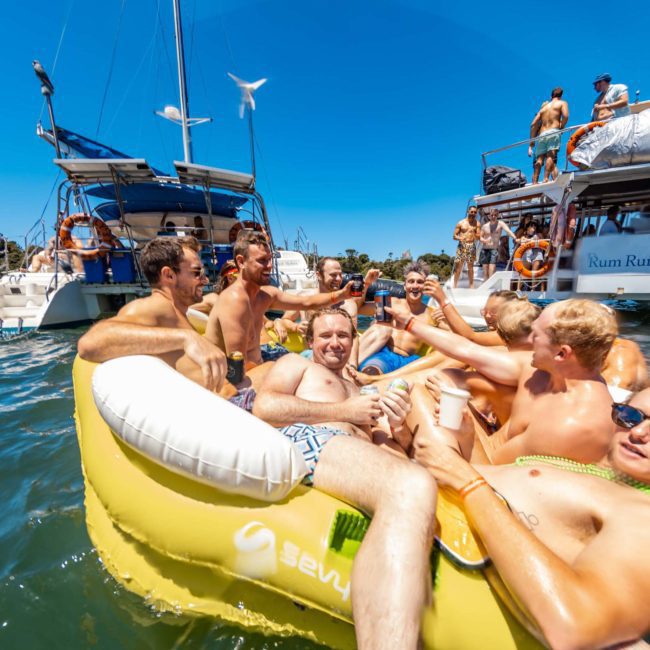 A group of people in swimwear enjoying a sunny day on inflatable flotation devices near anchored boats on the water, with a luxury yacht hire Sydney adding to the festive atmosphere.