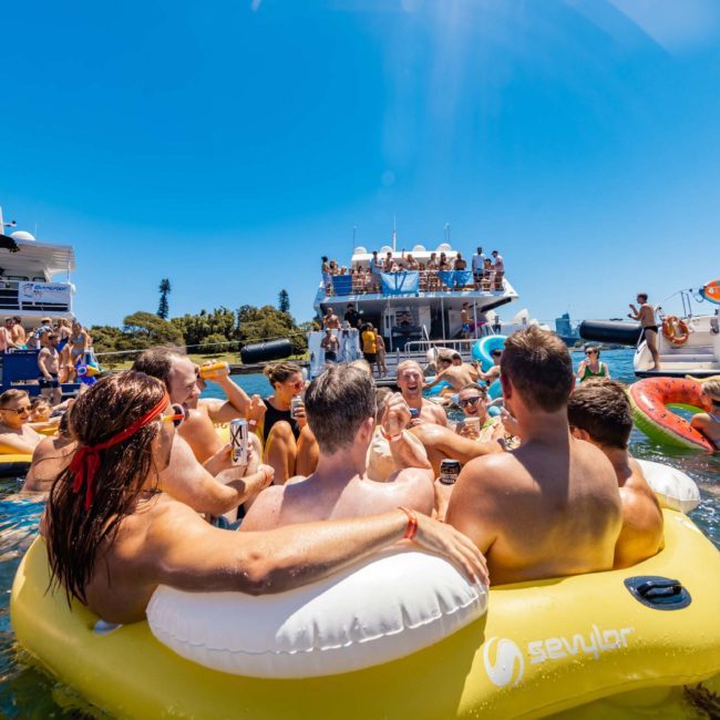 People enjoying a summer party on inflatable rafts and boats in a sunny waterfront setting with buildings and trees in the background, featuring Sydney boat party hire for an unforgettable experience.