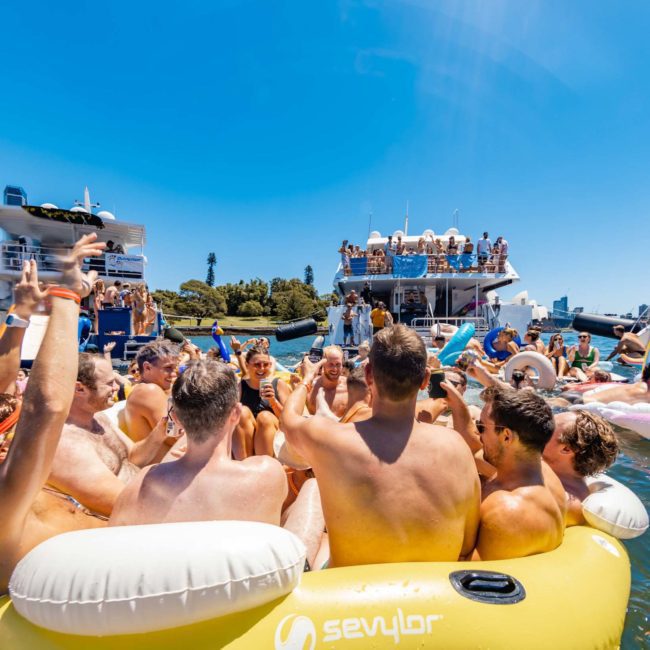 A large group of people in swimsuits are gathered on inflatable floats in the water, raising drinks. Boats with more people in swimsuits are docked nearby against a backdrop of tall buildings and blue sky, enjoying the atmosphere created by a DJ boat hire Sydney.