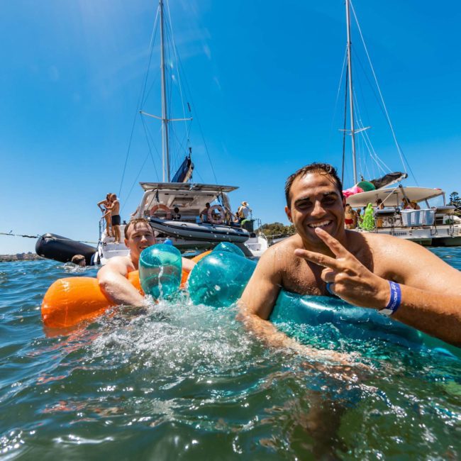 A man in a pool on an inflatable float gives a peace sign while people and boats, including luxury yacht hire Sydney, are seen in the background under a clear blue sky.
