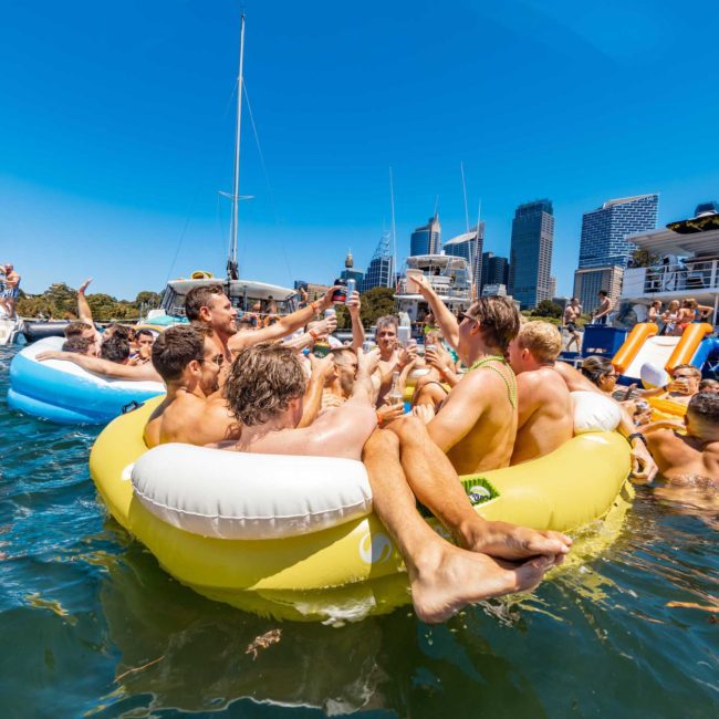A group of people in swimwear are gathered on an inflatable raft in the water surrounded by boats and a city skyline, enjoying what looks like a fun Sydney boat party hire.