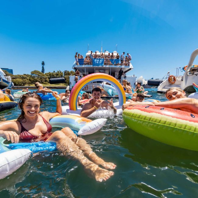 Adults enjoying a sunny day floating on colorful inflatables in the water, with luxury yachts and boats in the background. Some people are on board, soaking up the clear skies. Perfect for a Sydney boat party hire or DJ boat hire Sydney experience.