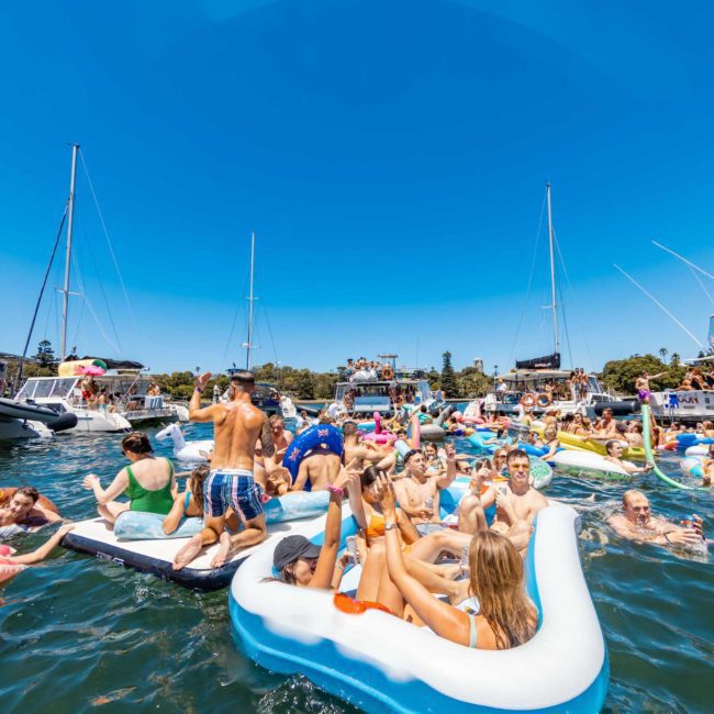A large group of people are enjoying a sunny day on the water, relaxing in and around inflatable floats and boats. For an elevated experience, consider a luxury yacht hire Sydney or private yacht charter on Sydney Harbour.