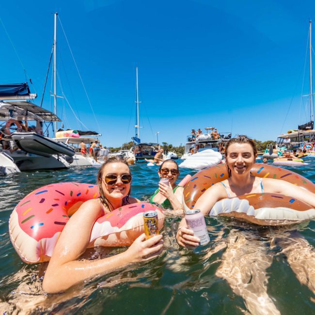Three people in donut-shaped floaties holding beverage cans, smiling in a lively water area with boats and other people in the background on a sunny day, enjoying the vibrant atmosphere of a DJ boat hire Sydney.
