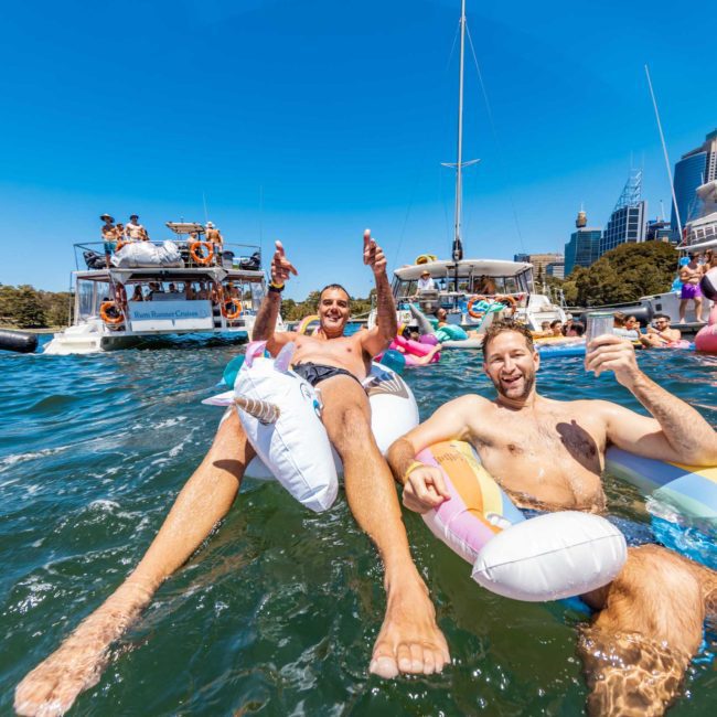 Two people are floating on inflatable loungers in a body of water during a catamaran party in Sydney, surrounded by boats and other people. One person is holding a drink, and both are smiling and gesturing towards the camera.
