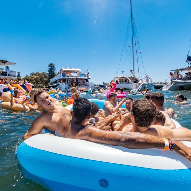A group of people relax on inflatable rafts in a crowded bay with boats in the background under a clear blue sky, enjoying what seems like a perfect day for a catamaran party in Sydney.