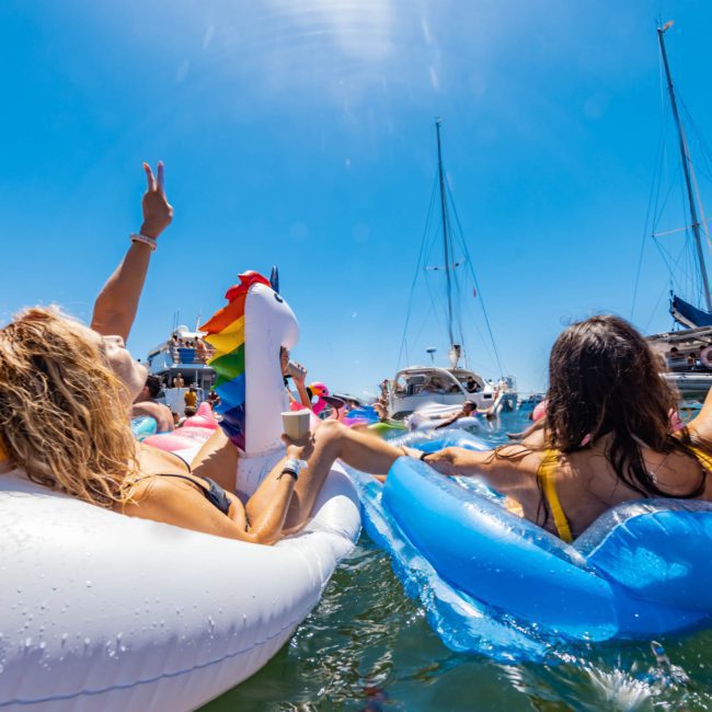 People on colorful inflatable pool floats enjoy the water, with sailboats and a bright blue sky in the background. Imagine adding to the experience with a private yacht charter Sydney Harbour or DJ boat hire Sydney for an unforgettable day.