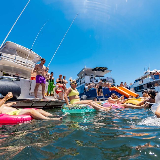 People in swimsuits are lounging on inflatable floats near a docked yacht and boats on a sunny day. The water is calm, and several boats can be seen in the background, perfect for a DJ boat hire Sydney or a lively catamaran party Sydney.