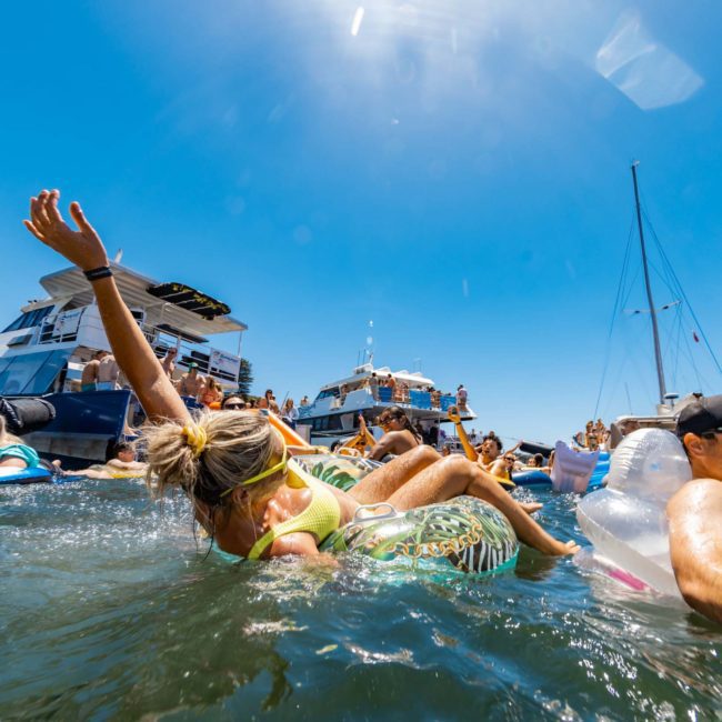 People are floating on inflatable tubes and relaxing in the water near several boats on a sunny day, enjoying a lively atmosphere thanks to a DJ boat hire Sydney.