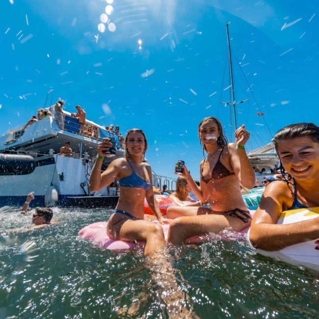 A group of people enjoying a sunny day on a luxury yacht hire in Sydney and in the water, with three women in bikinis holding drinks while sitting on floating devices.
