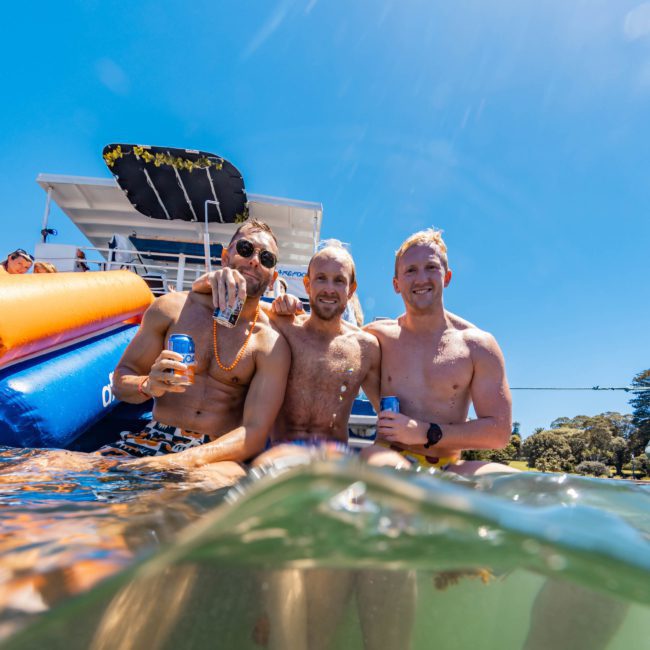 Three shirtless men are standing in the water near a boat, holding canned drinks, and smiling at the camera on a sunny day during a catamaran party in Sydney.