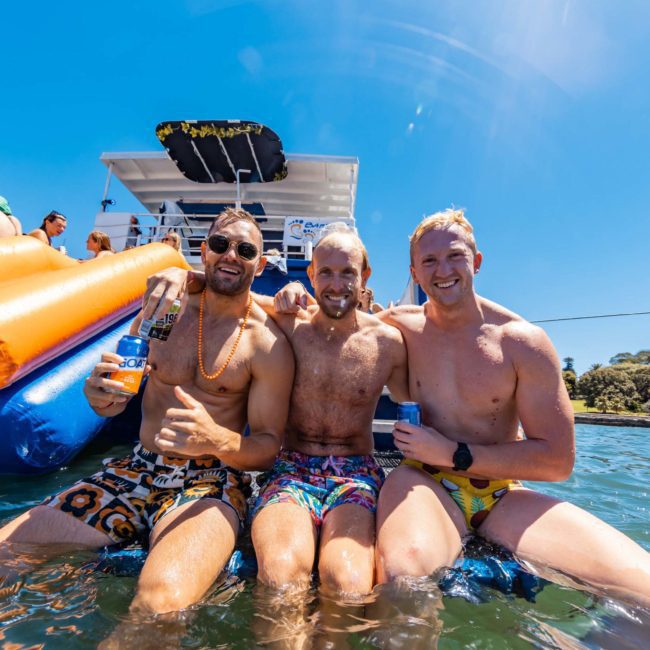 Three men in colorful swim trunks sit on an inflatable slide in the water, holding drinks, with boats and a sunny blue sky in the background. They are smiling and appear to be enjoying a sunny day at a Sydney boat party hire.