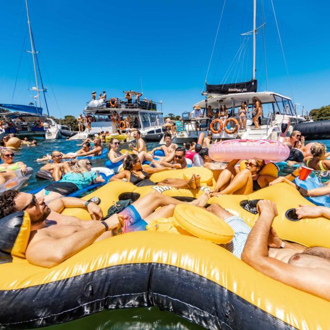 A group of people floats on inflatable rafts and inner tubes in the water near boats on a sunny day, enjoying a festive Catamaran party in Sydney Harbour.
