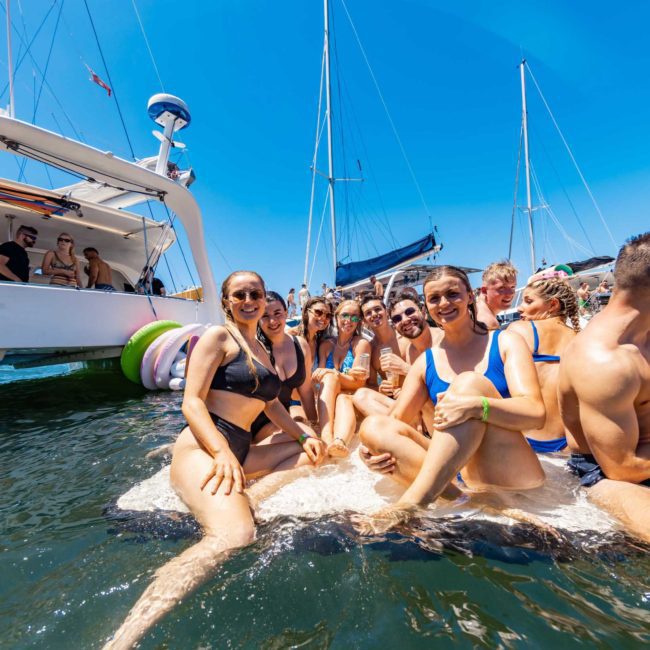 A group of people wearing swimwear sits and stands on a floating platform in the water near a catamaran, under a clear blue sky, enjoying a private yacht charter in Sydney Harbour.