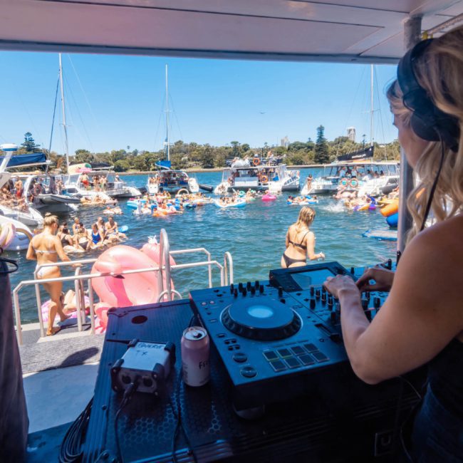 A DJ performs on a catamaran party in Sydney, facing a lively crowd of people on floaties and other boats in the water, with clear skies and trees in the background.