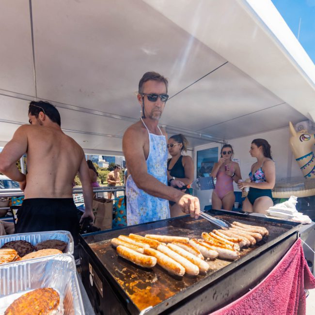At an outdoor event, a person in sunglasses and an apron cooks sausages on a large grill, with people in swimsuits nearby. In the background, you can spot a private yacht charter in Sydney Harbour. A tray of cooked patties is also visible on the side.