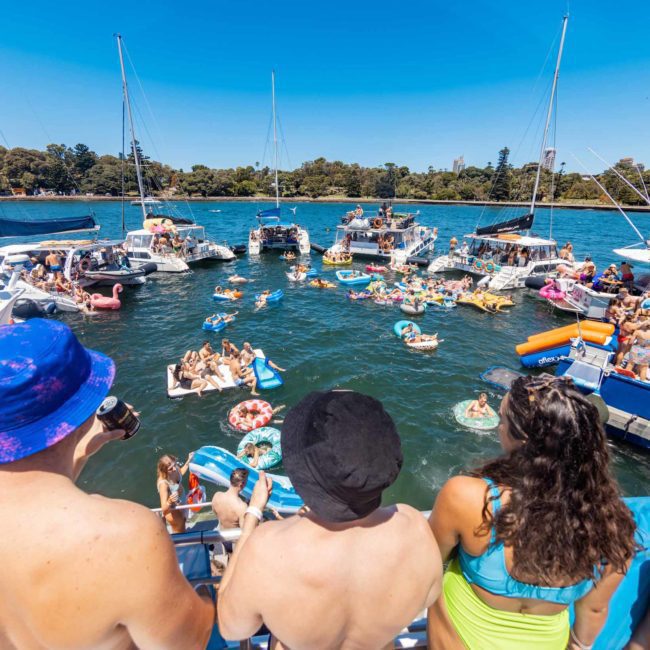 People gather on boats and inflatables on a sunny day in a lively body of water surrounded by trees, enjoying what feels like a private yacht charter on Sydney Harbour.
