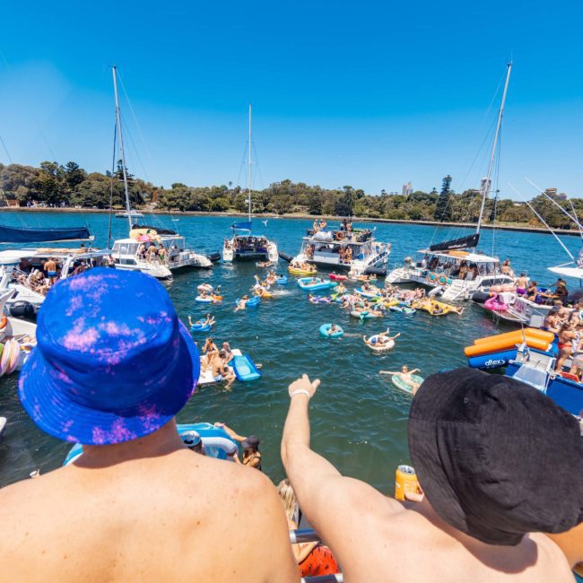 Two people wearing hats and swimsuits stand at the edge of a boat, looking at numerous boats and people swimming and lounging in a sunny water setting. Trees line the shore in the background, perfect for a Sydney boat party hire or a private yacht charter on Sydney Harbour.