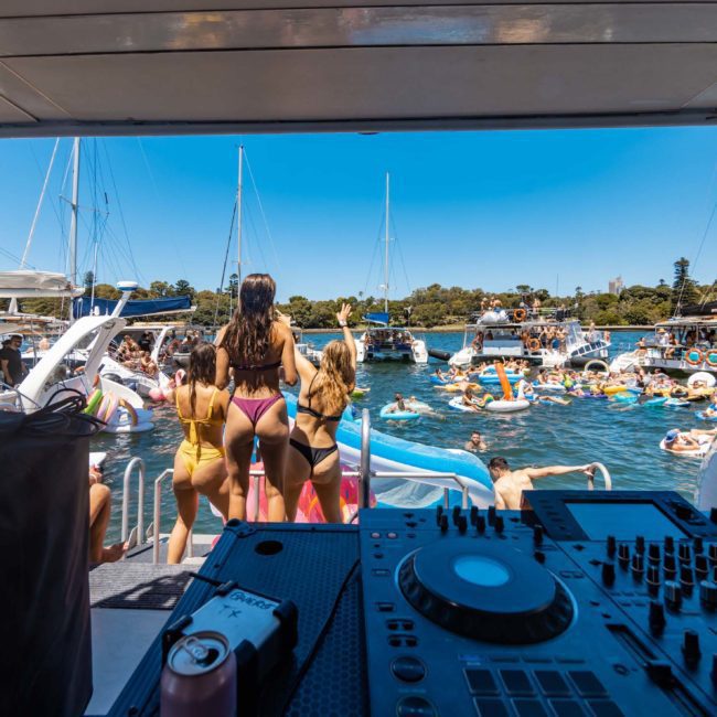A DJ is playing music on a boat while people in swimwear enjoy the water and other boats around them on a sunny day, making it the perfect Sydney boat party hire experience.