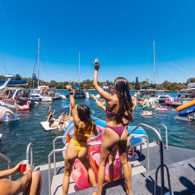 People in swimsuits enjoying a sunny day on boats and inflatables in a crowded water area, with a vibrant Catamaran party Sydney happening nearby.
