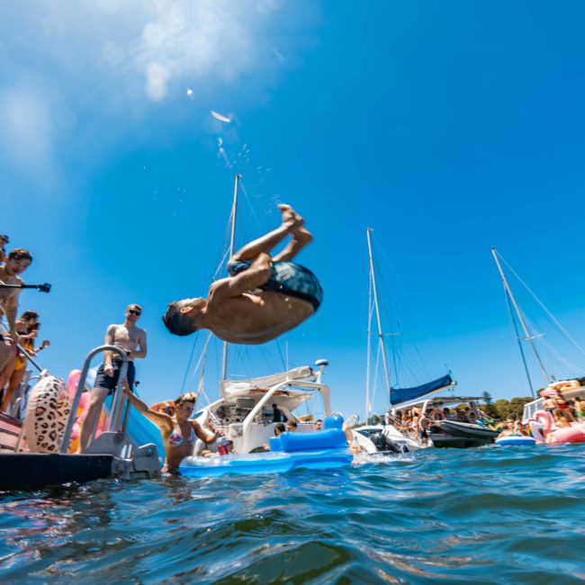 A person is mid-backflip off a boat into the water, surrounded by other boats and people enjoying the sunny day, epitomizing the excitement of a DJ boat hire Sydney event.